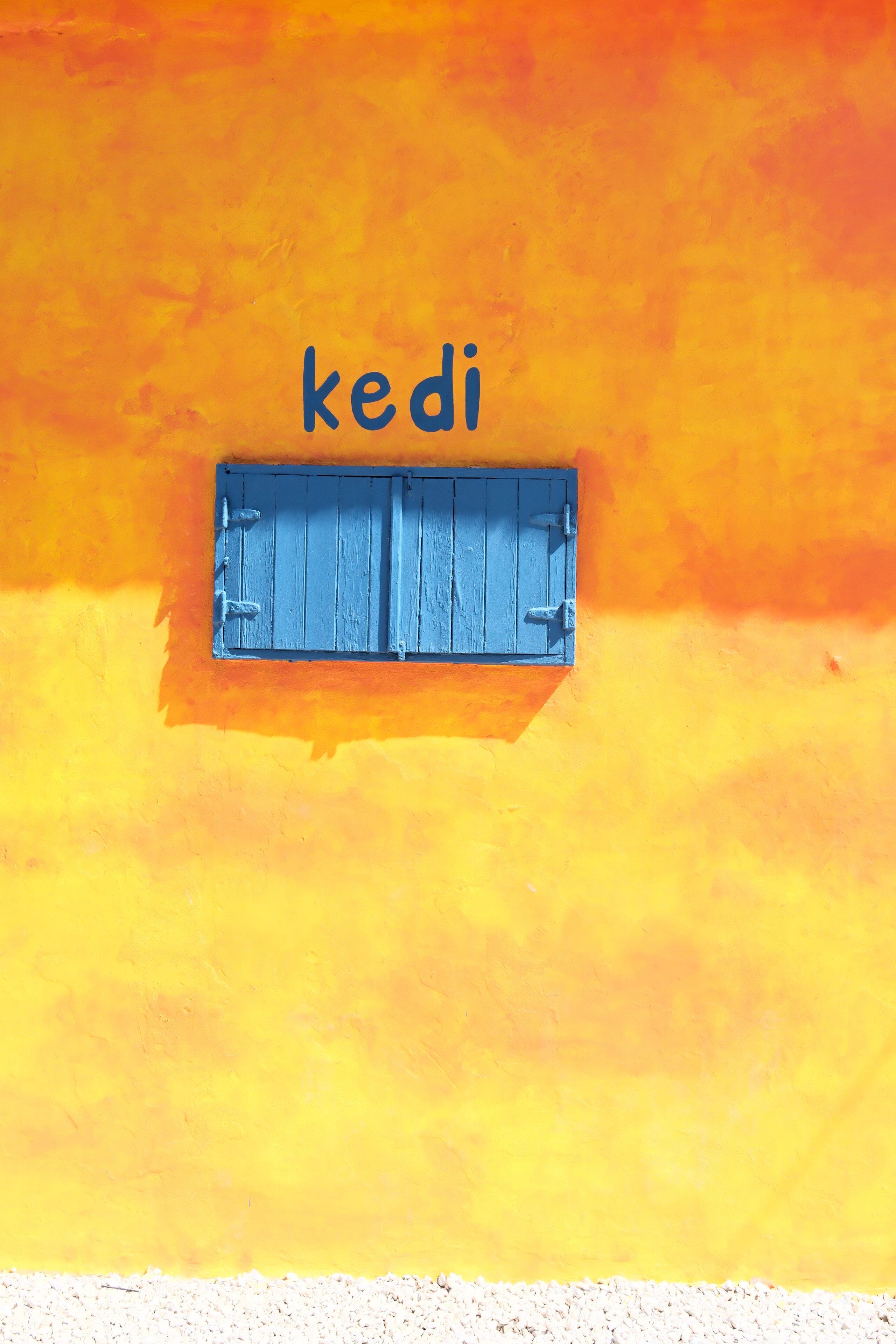 Bright yellow wall with a 'kedi' text and blue shuttered window. Minimalist and colorful.