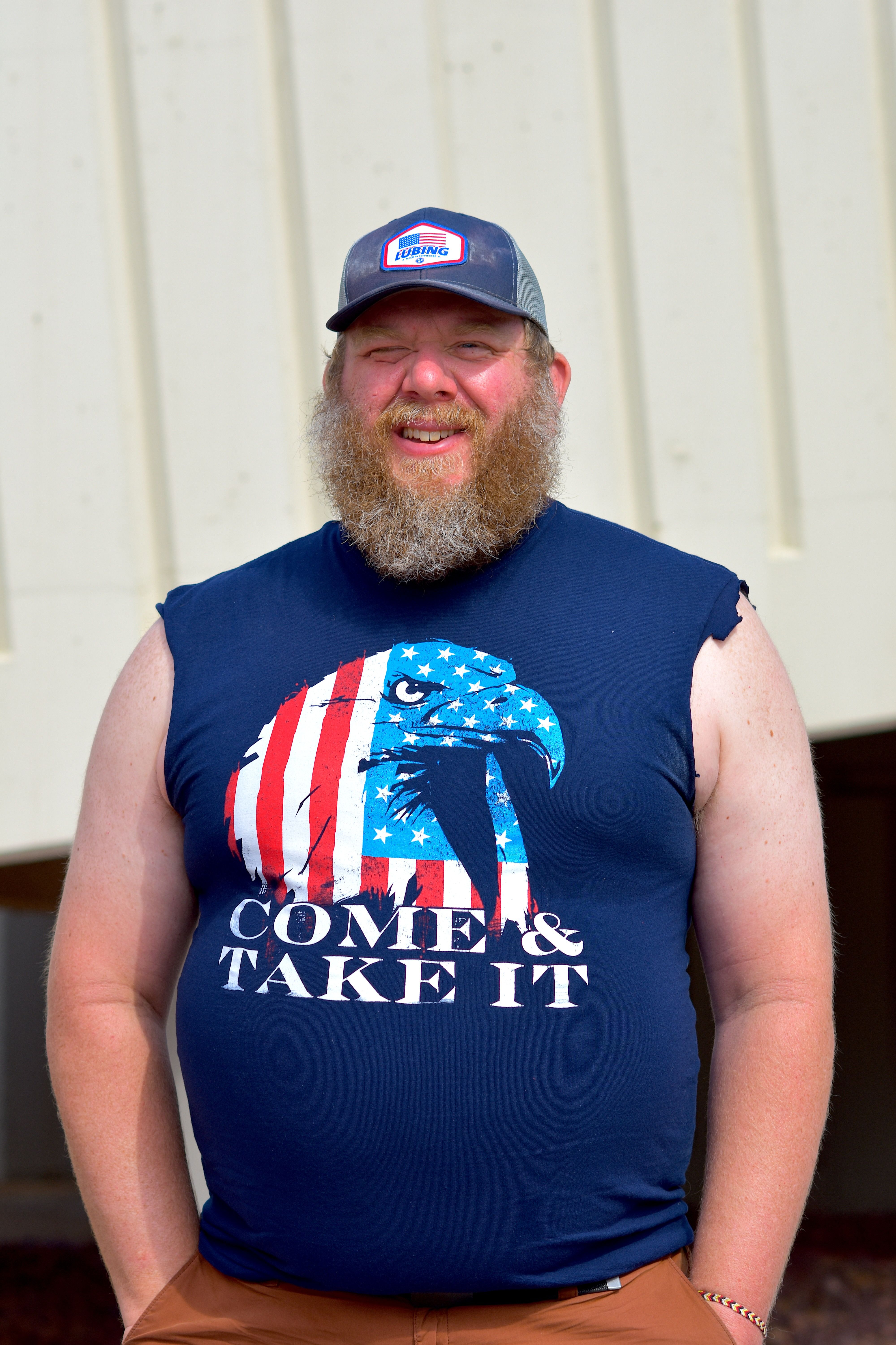 A bearded man with a cap smiles in a sleeveless patriotic T-shirt outdoors.