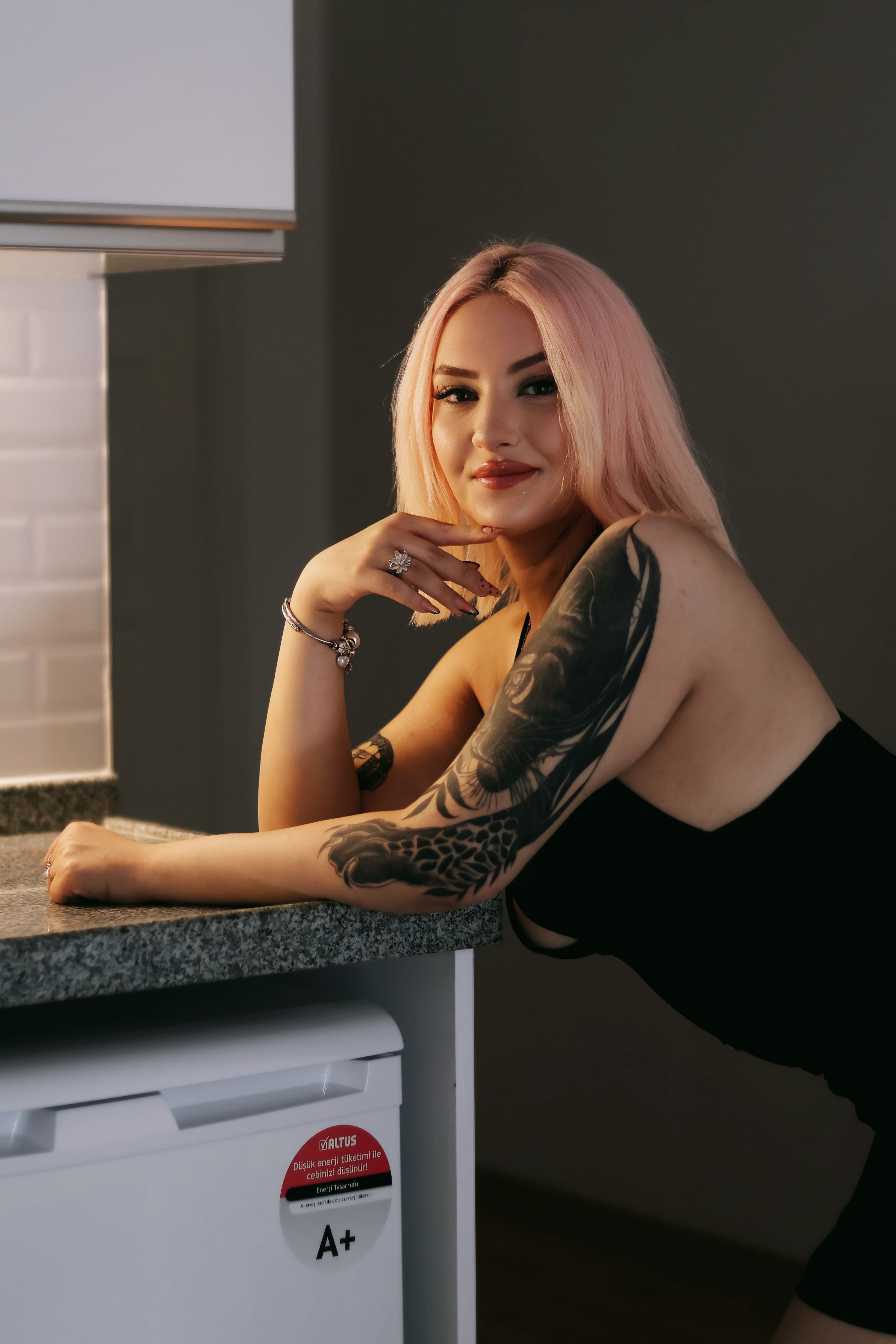 A young woman with pink hair and tattoos in a stylish indoor setting.