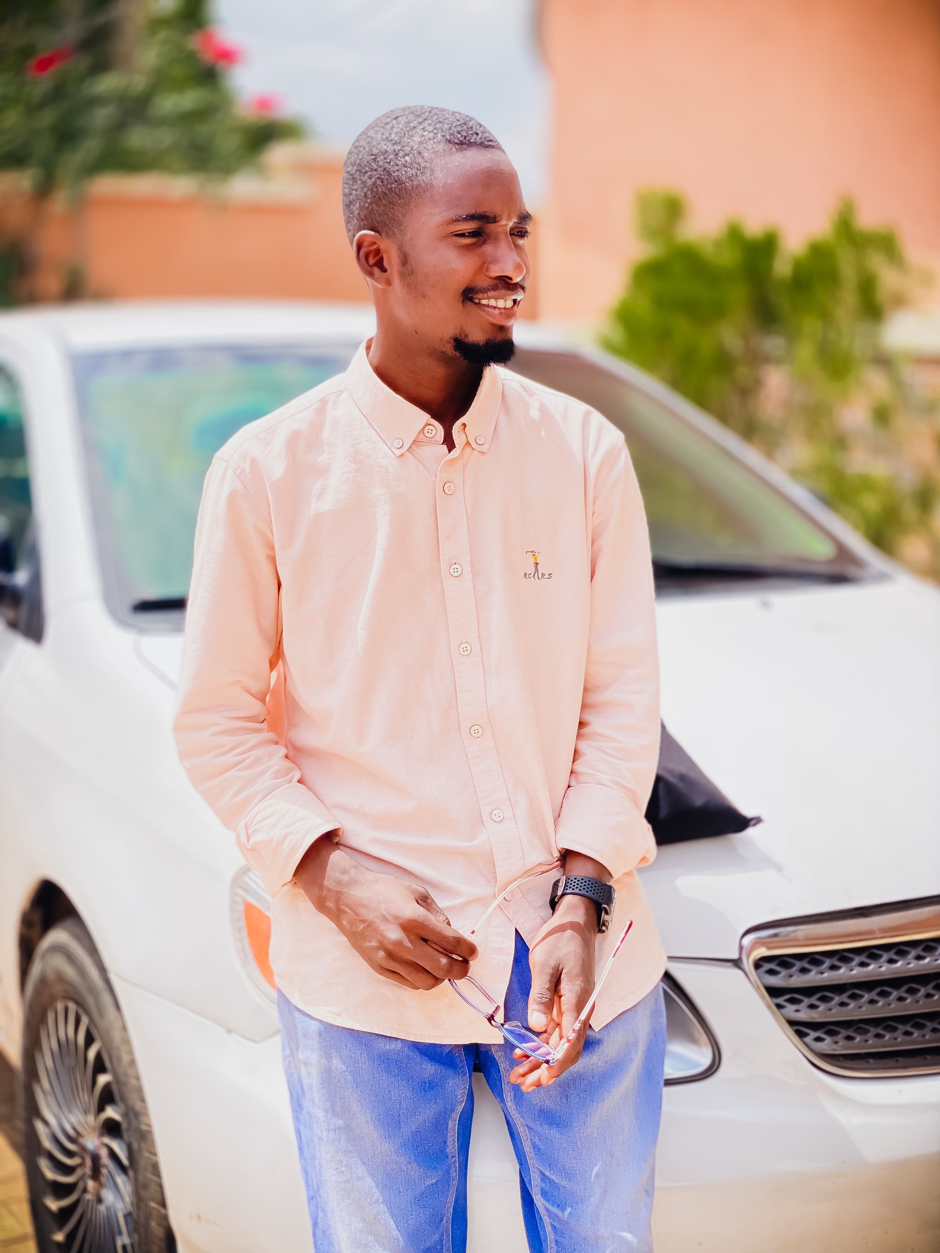 Smiling young adult male standing beside a parked car outdoors on a sunny day.