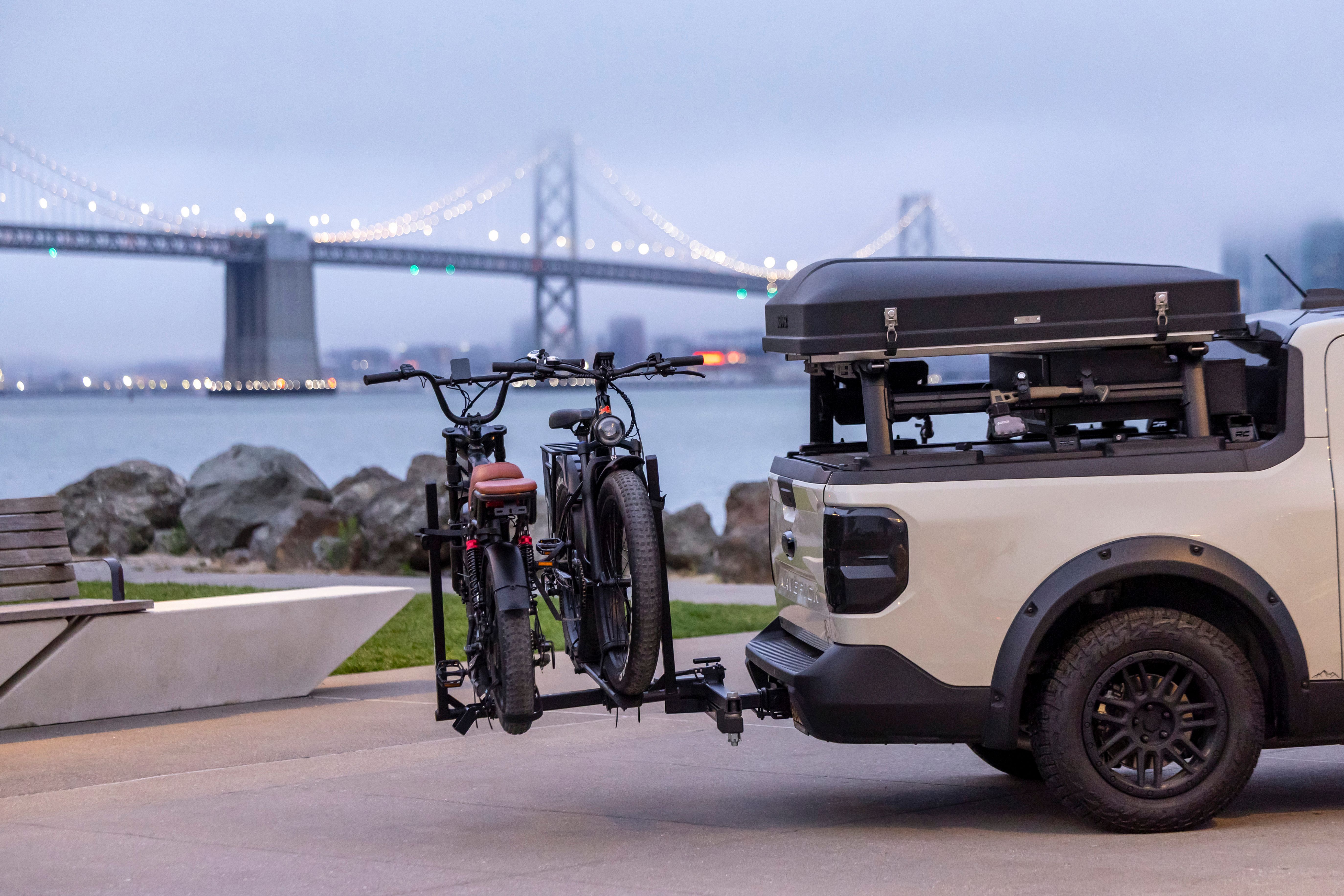 Electric pickup truck with two bicycles mounted on a rack, parked near the Bay Bridge at twilight.
