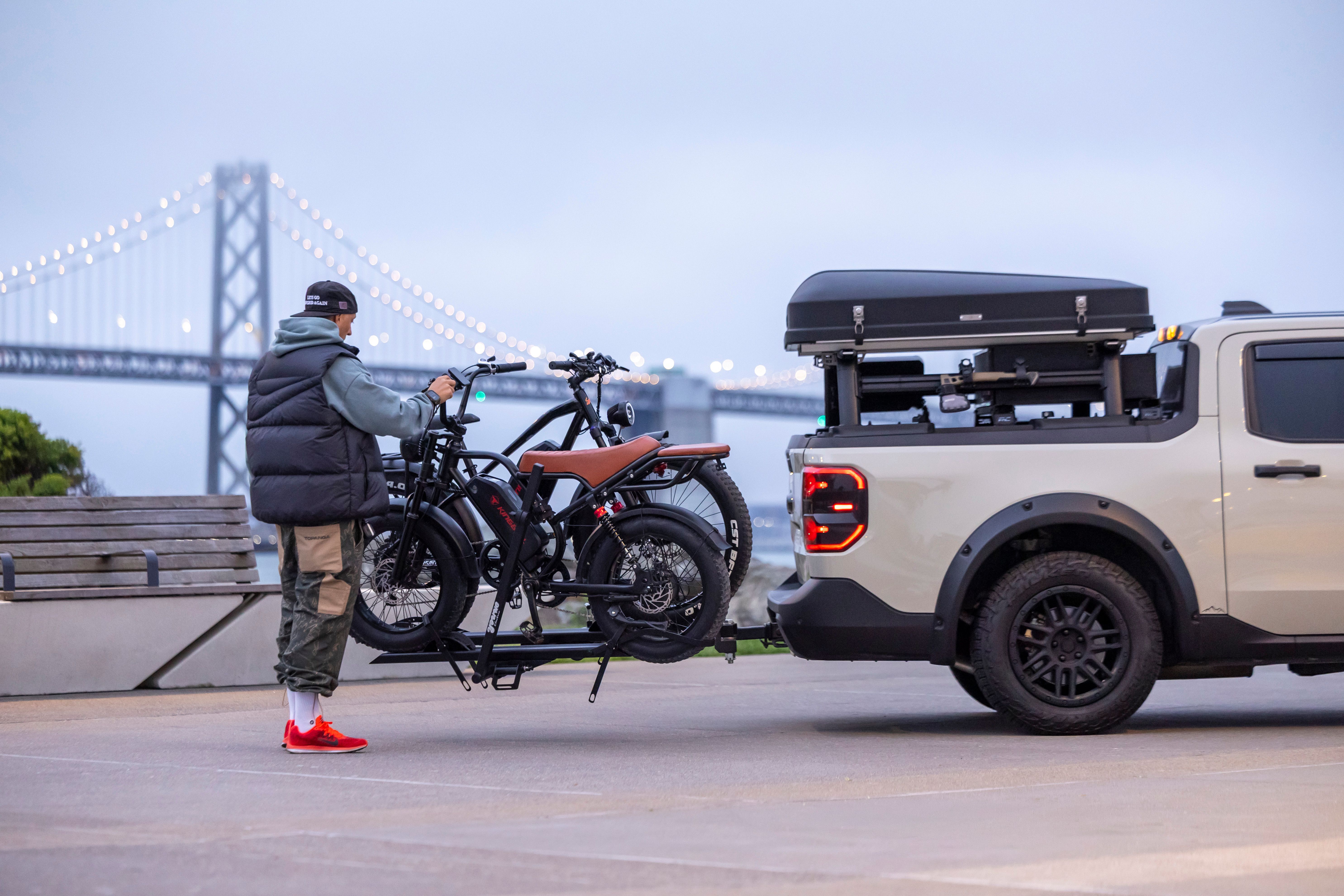 Man loading electric bikes onto a truck near a large suspension bridge.