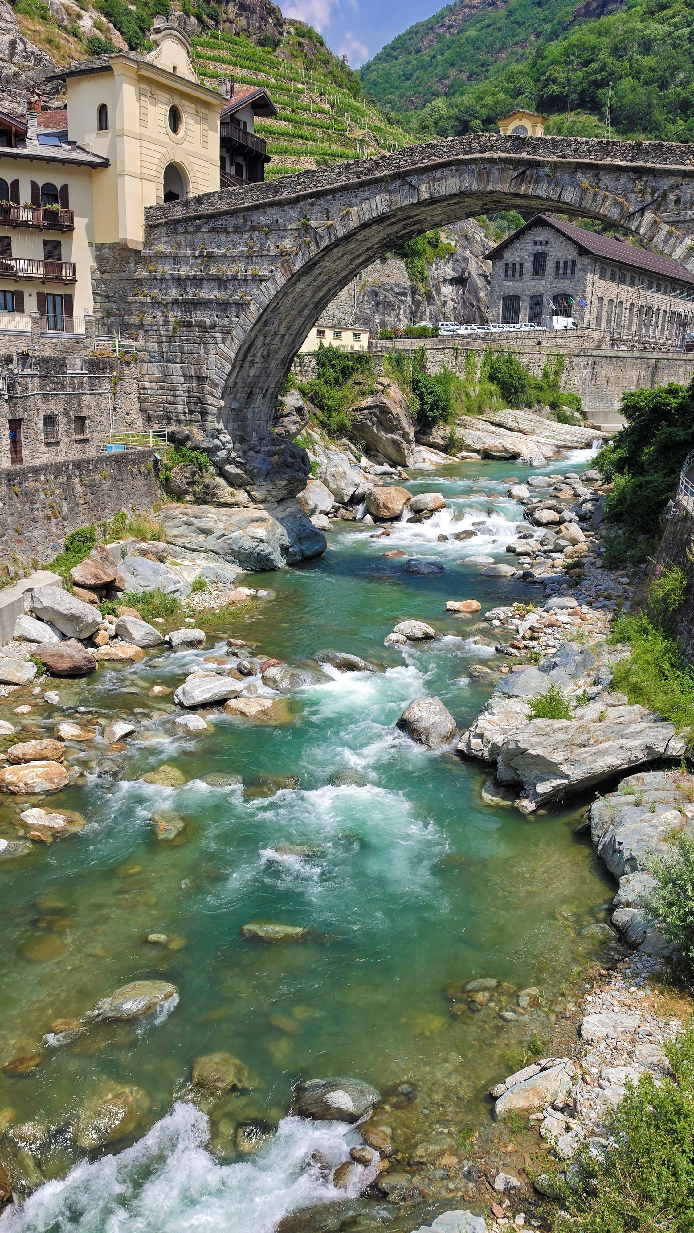 Scenic view of a historic stone bridge over a clear mountain river in Italy's Aosta Valley.