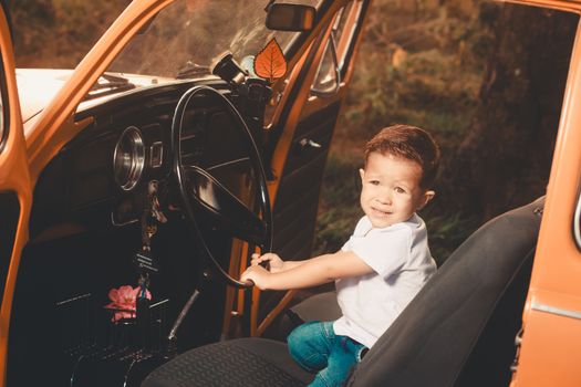 Cute child sitting in an orange vintage car, pretending to drive while looking at the camera.