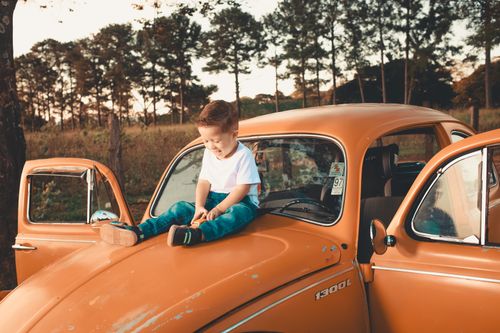 Free A joyful child sitting on the hood of an orange vintage car in a sunny park setting. Stock Photo