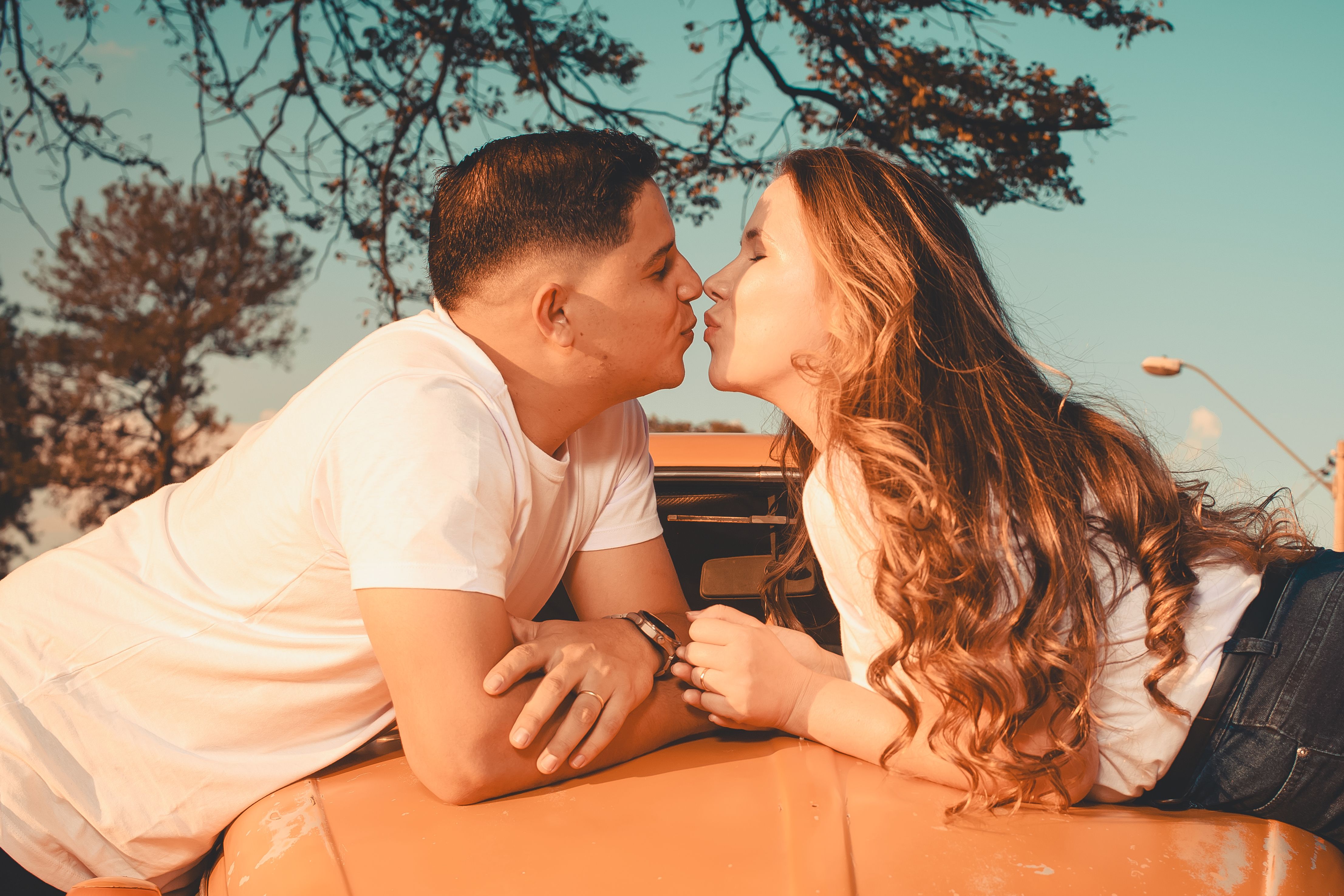Playful couple kissing while leaning on a car in a warm, sunny outdoor setting.