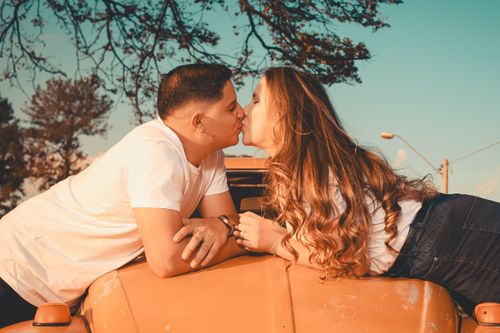 Free Romantic young couple sharing a kiss on a vintage car during a sunny day, surrounded by trees. Stock Photo