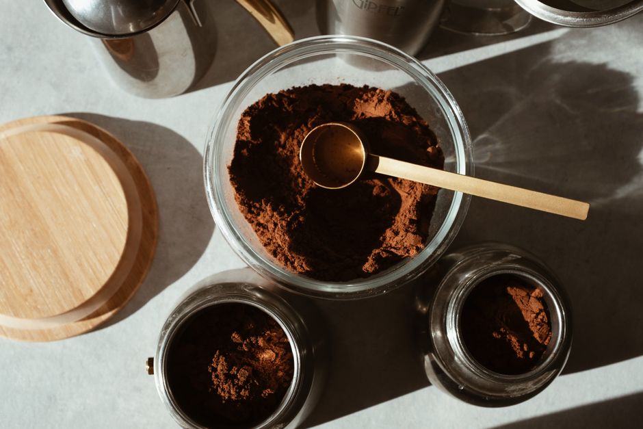 A glass jar filled with dark chocolate powder mix next to a wooden scoop and a mug of hot chocolate made from it.