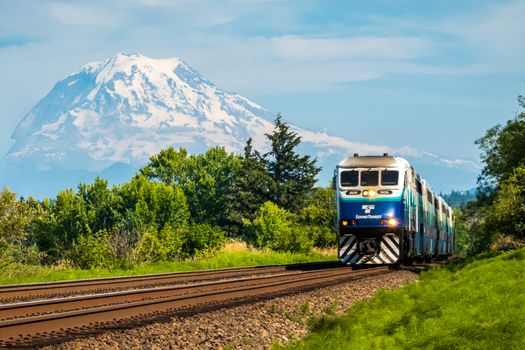 Passenger train traveling with Mount Rainier in the background, lush greenery, bright skies.