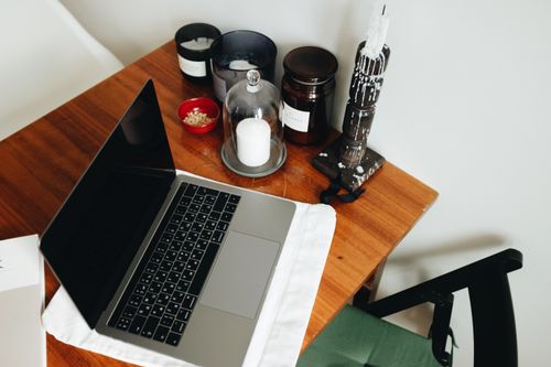 Free A home office with a laptop on a wooden table, surrounded by candles and jars for a warm atmosphere. Stock Photo