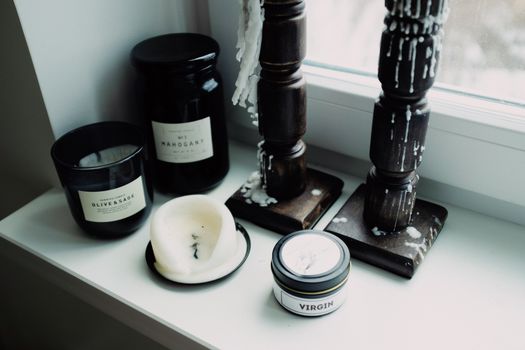 High angle of various candles placed in black glass jars and wooden candlestick arranged on white surface of windowsill in light room