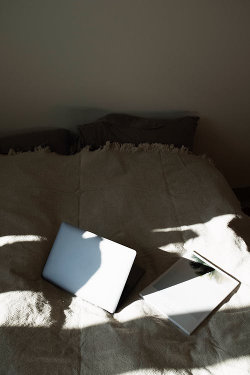 Top view of laptop and book placed on soft blanket on comfortable bed in morning light in modern bedroom