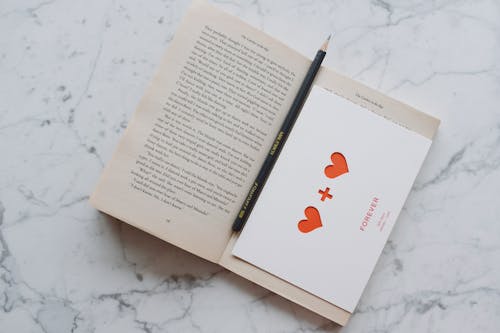 Top view of opened novel book with pencil and romantic postcard placed on white marble surface