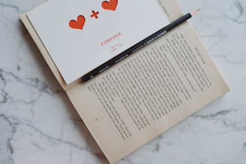 Top view of opened novel book with pencil and romantic postcard with hearts placed on white marble surface