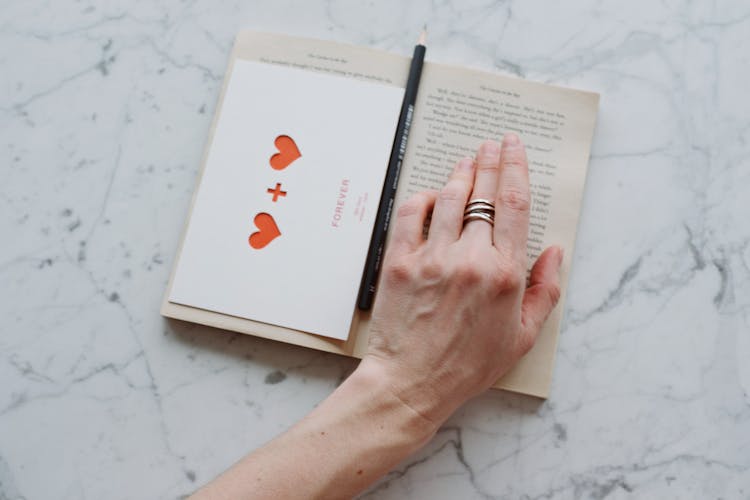 Anonymous Crop Person Reading Book With Postcard And Pencil