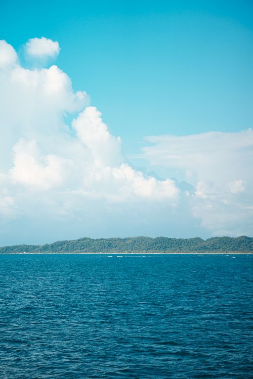 Free Calm blue ocean with distant island and fluffy clouds under a clear sky, ideal for stress relief themes. Stock Photo
