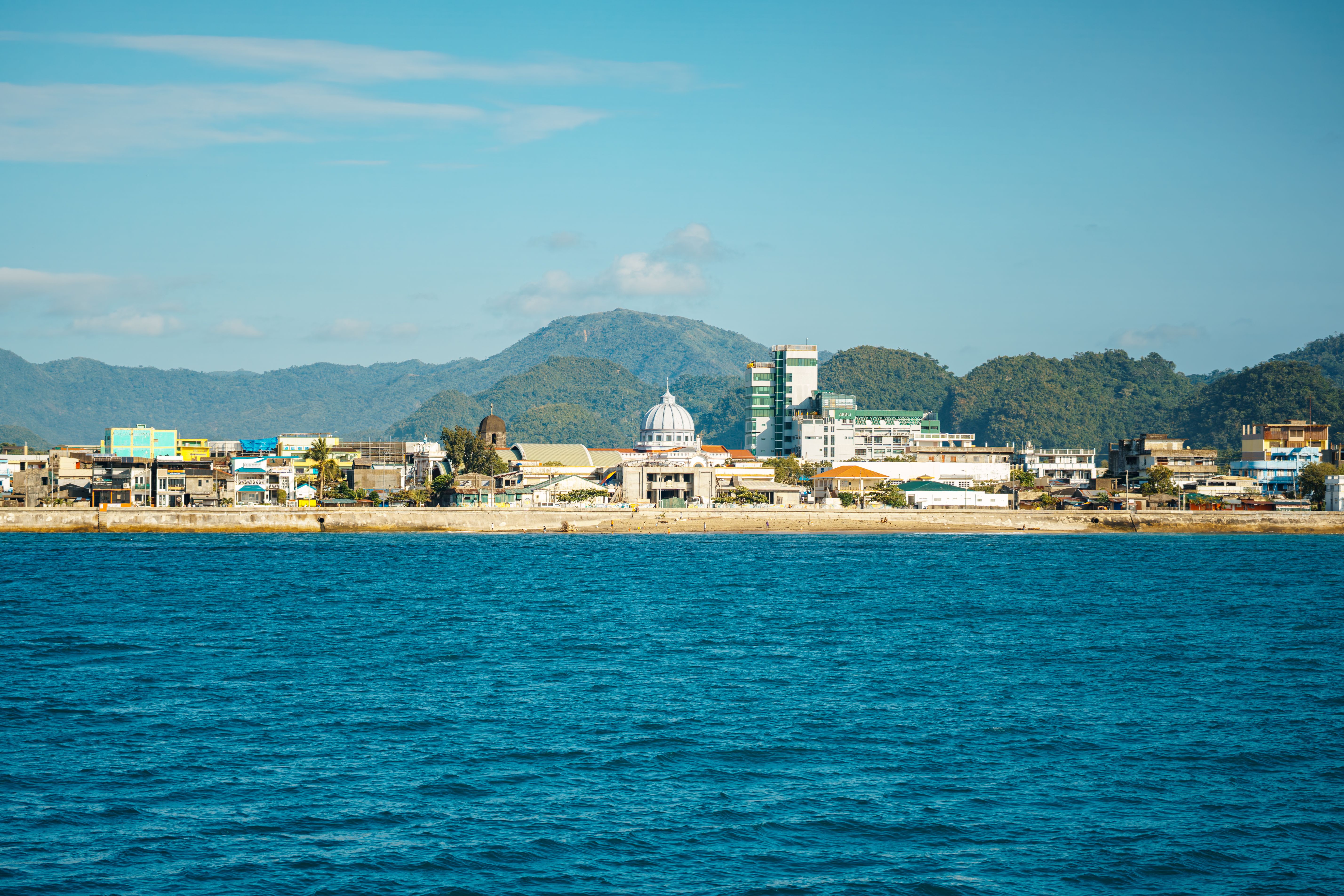 Beautiful view of a coastal city against a backdrop of lush mountains, under a clear blue sky.