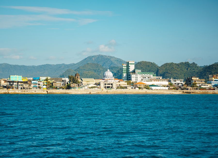 Beautiful view of a coastal city against a backdrop of lush mountains, under a clear blue sky.