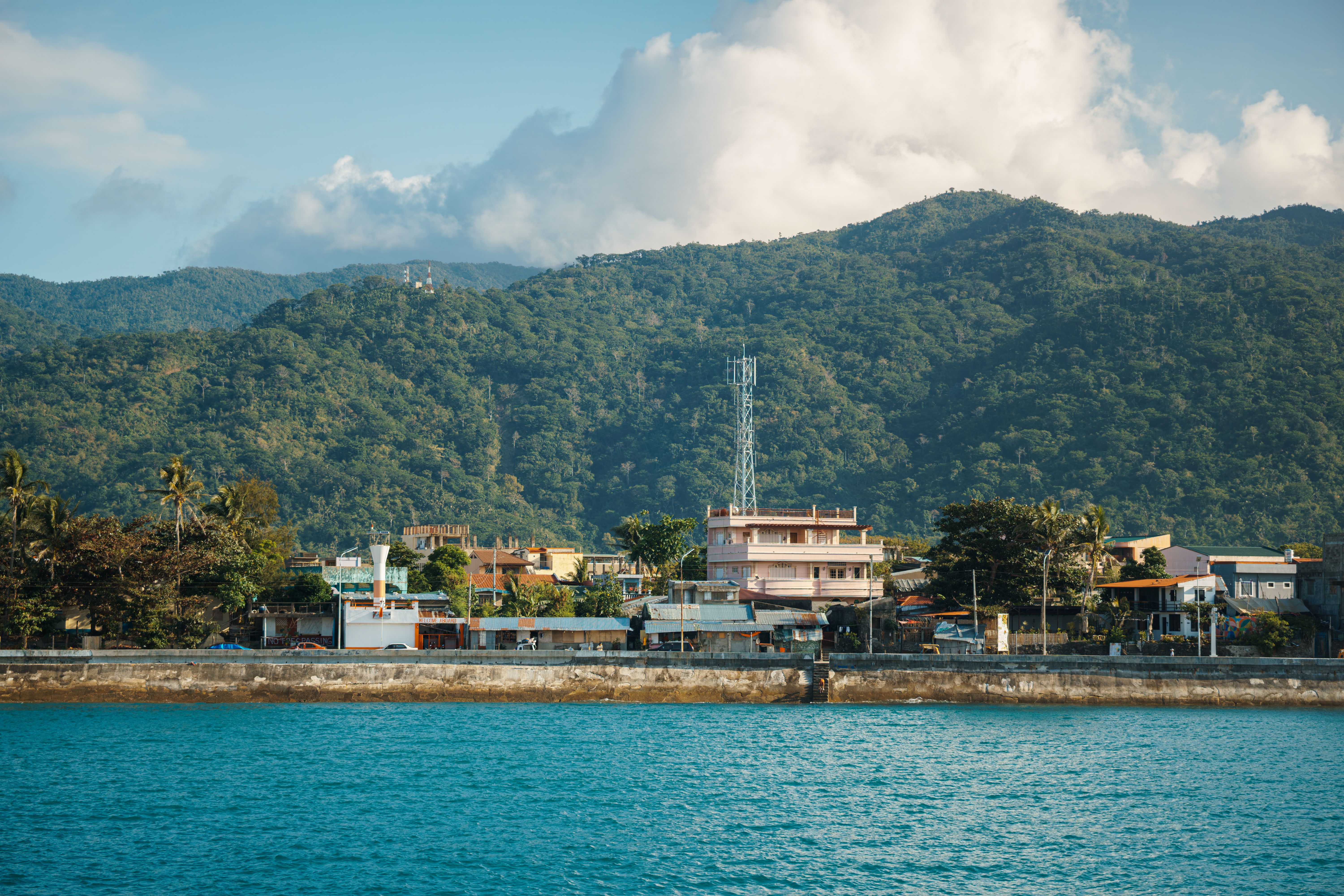 A Picturesque Coastal Village With Lush Mountains In The Background On A Sunny Day.