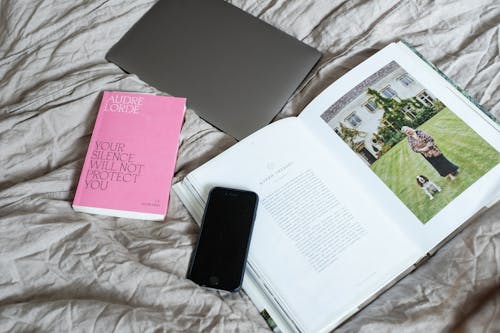 Top view of arranged art book with smartphone and laptop placed on soft blanket on comfortable bed in modern light room