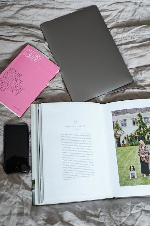 From above view of opened book with smartphone and laptop placed on soft blanket on comfortable bed in modern light bedroom