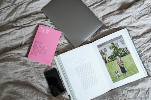 Top view of opened book with laptop and smartphone placed on soft blanket on comfortable bed in modern light bedroom