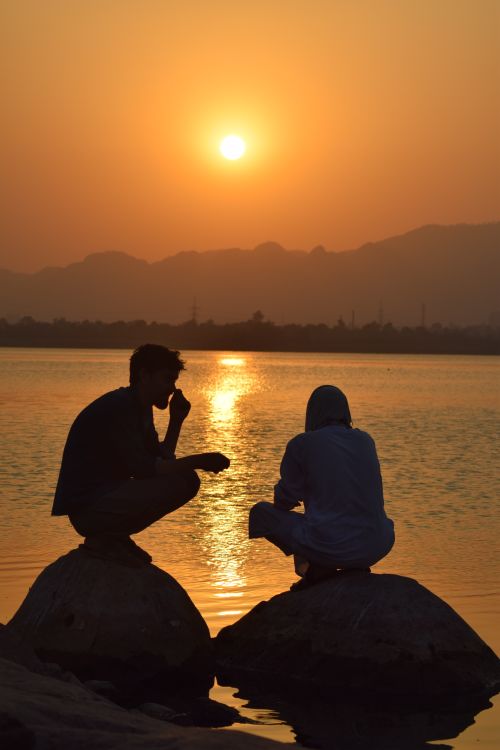 Free Two people sitting by the lake at sunset in Islamabad, creating a serene silhouette scene. Stock Photo