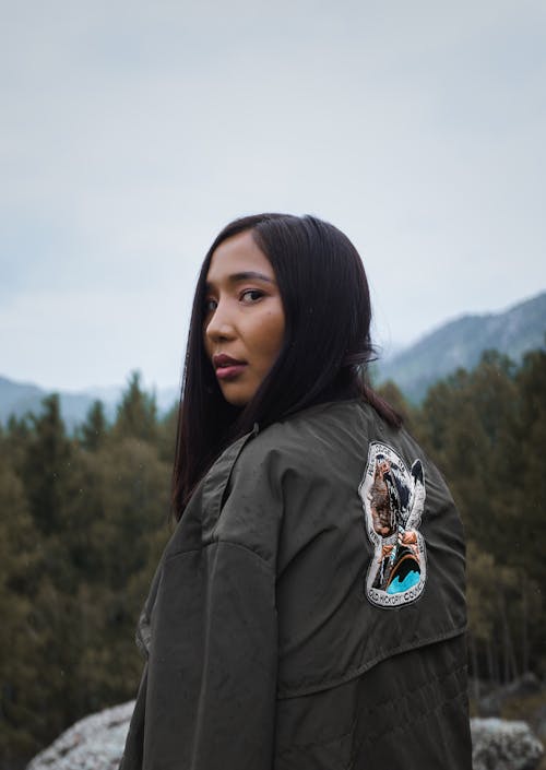 Back view of young ethnic lady with makeup in jacket with bright trendy pattern standing near rough stone and mounts with trees under sky and looking at camera over shoulder