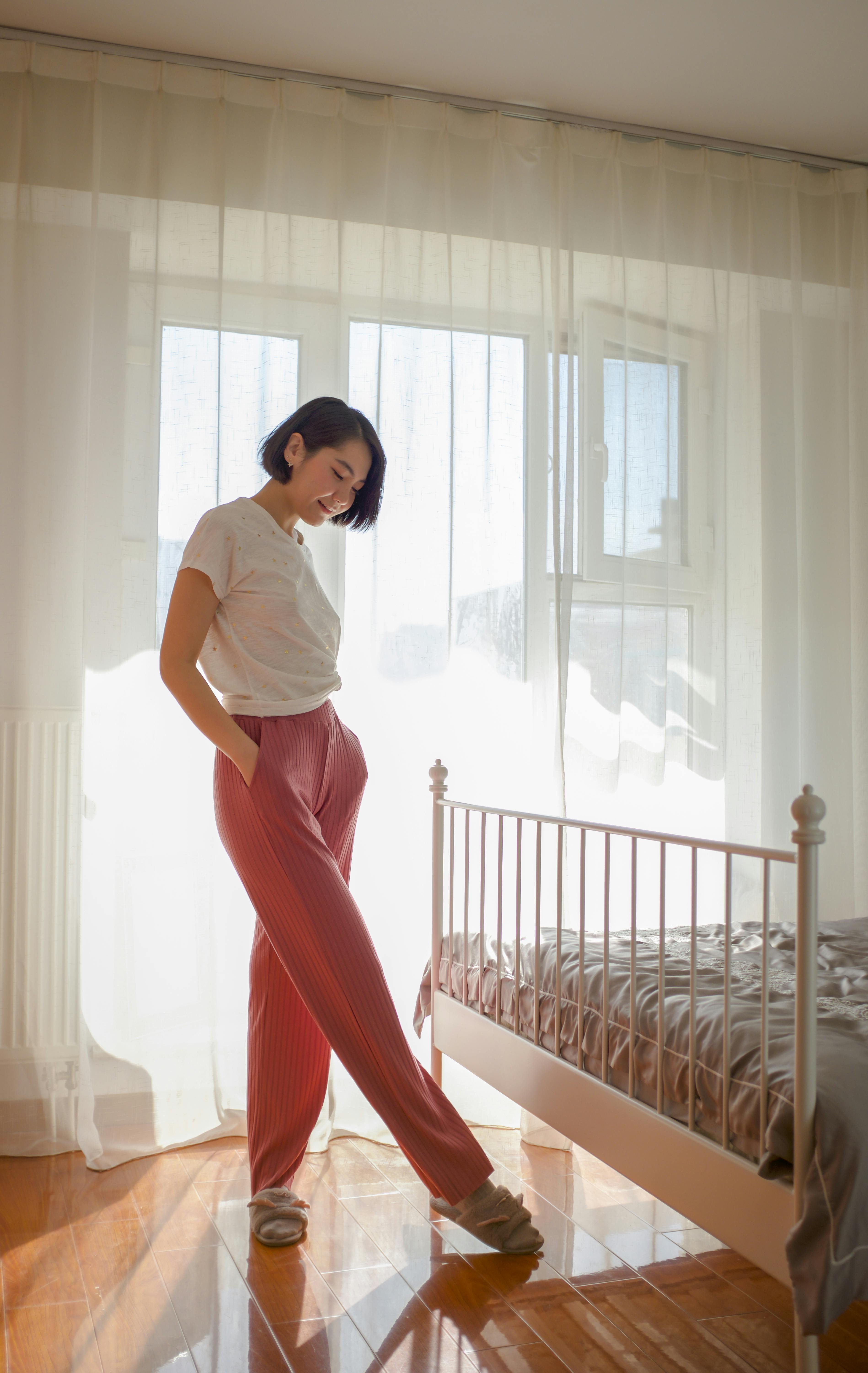 Free Side view of young slender ethnic lady in lounge wear and soft slippers standing with hand in pocket near bed and window with curtain while looking down at wooden floor Stock Photo