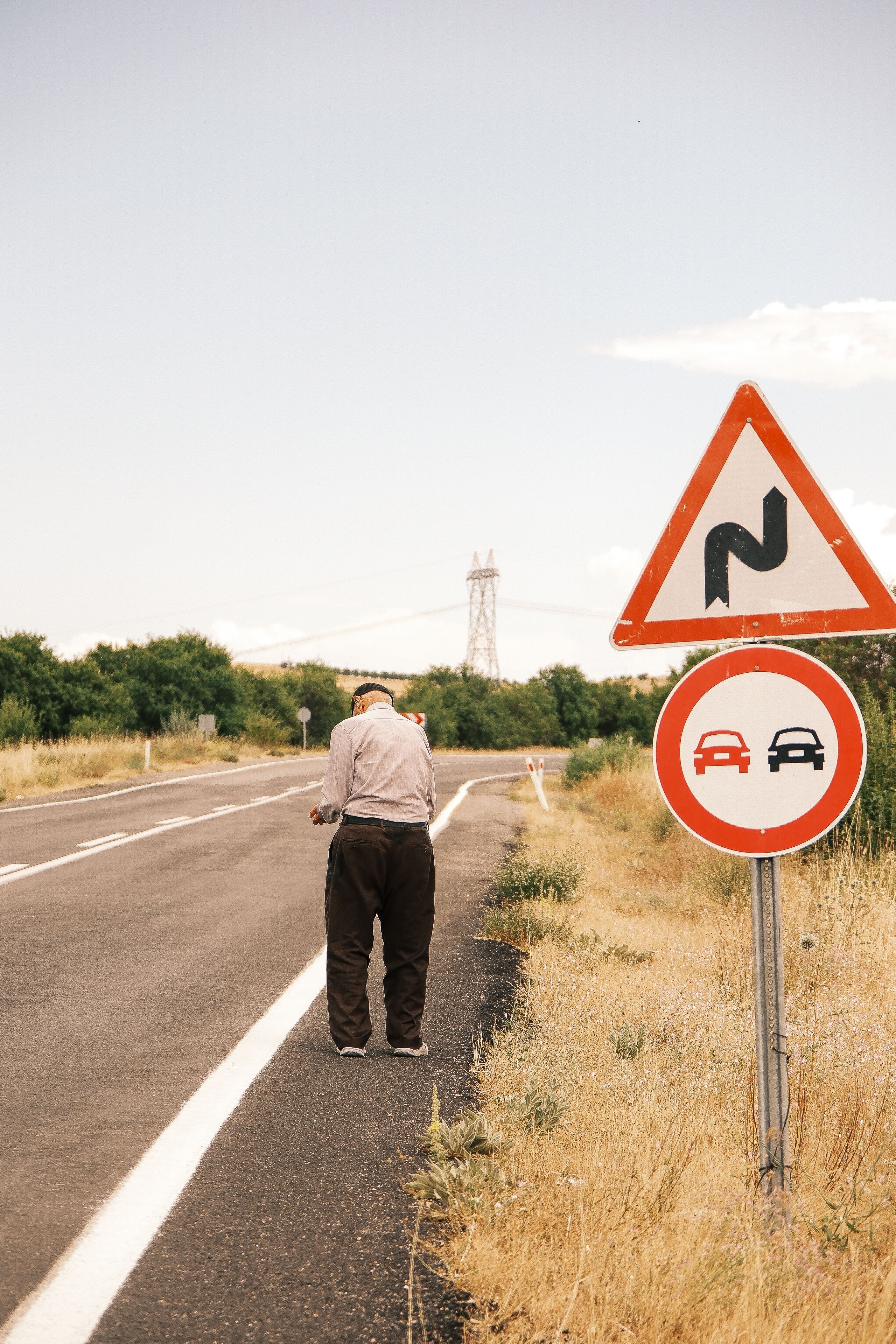 An elderly man walks along a rural road beside traffic signs in Türkiye. Daytime scene with nature.