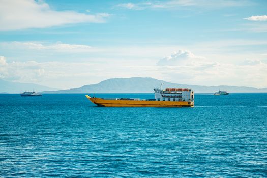 A yellow cargo ship sails through open blue waters under a clear sky, with distant land in view.