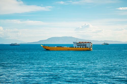 Free A yellow cargo ship sails through open blue waters under a clear sky, with distant land in view. Stock Photo