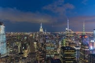 从现代城市上空俯瞰，夜间多云的天幕下，高层建筑的尖顶被人造光照亮