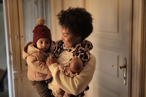 Cheerful ethnic woman hugging little kid while standing in room with toy and looking at clothes of baby while preparing to walk