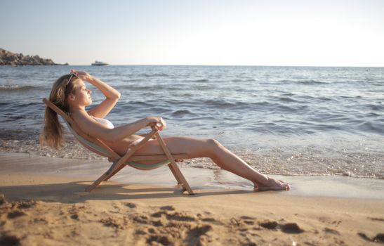 A woman sunbathing on a beach in a wooden chair by the ocean at sunset.