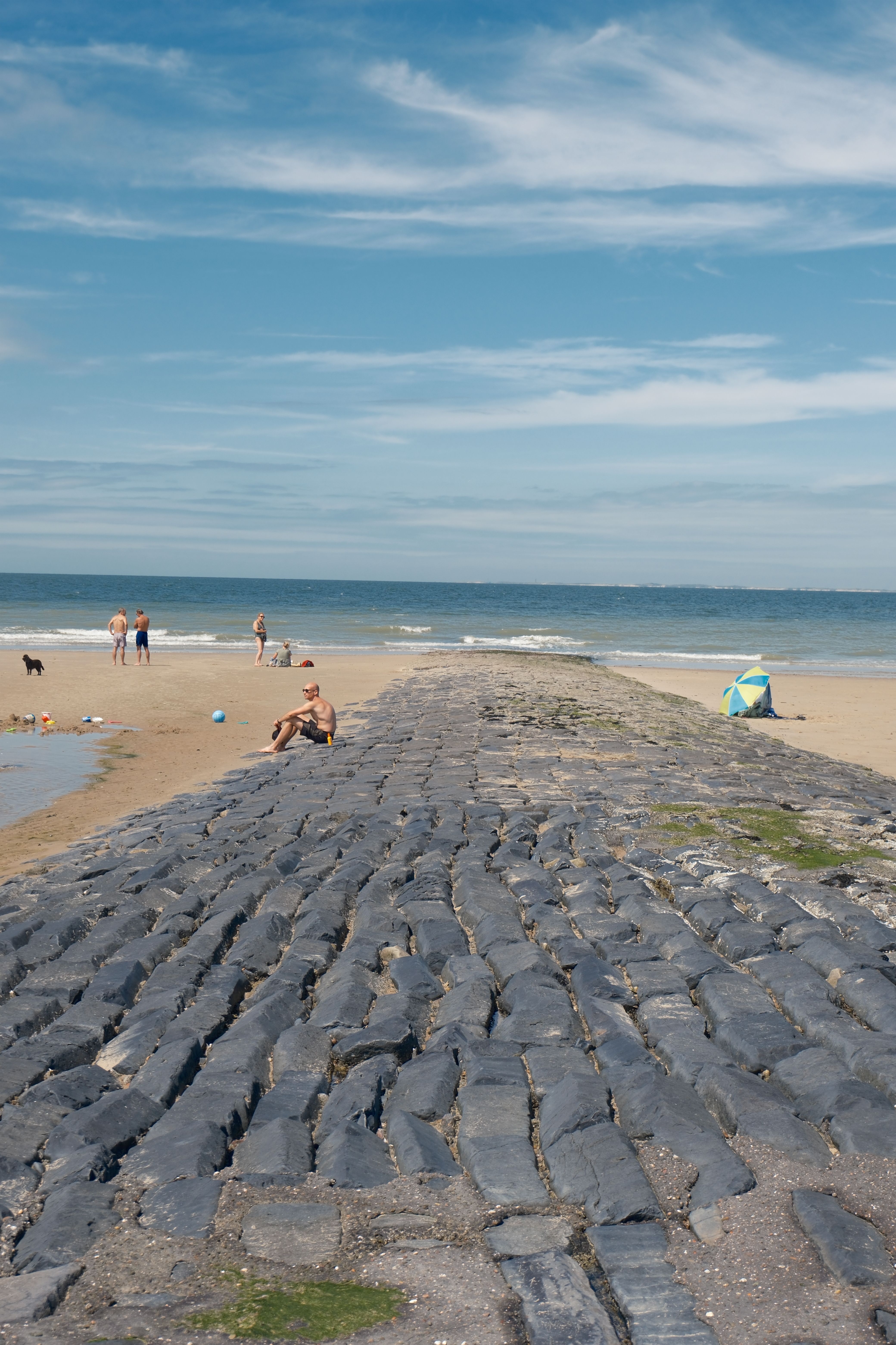 Sunny day at a beach with visitors relaxing on a stone jetty and sandy shore.