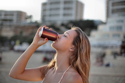 A woman sipping a drink from a can at the beach during sunset.
