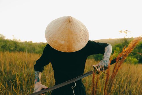 Free A tattooed individual wearing a conical hat with a samurai sword in a grassy outdoor setting. Stock Photo
