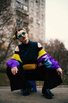 Low angle of stylish anonymous young female in colorful outfit and bunch hairstyle squatting on asphalt pavement near multistory building and trees in town