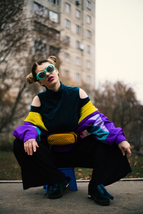 Free Low angle of stylish anonymous young female in colorful outfit and bunch hairstyle squatting on asphalt pavement near multistory building and trees in town Stock Photo