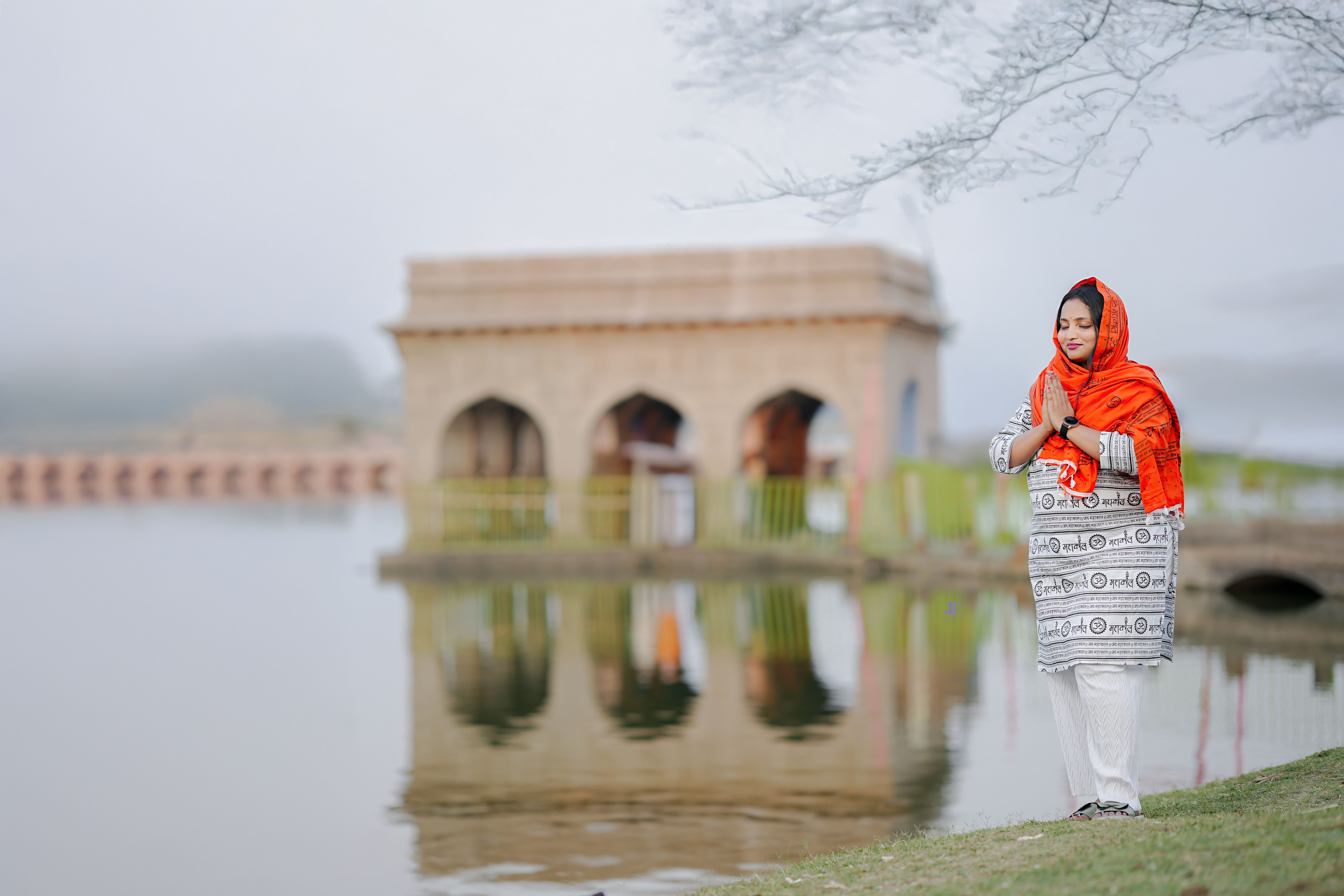 A woman in vibrant traditional attire stands in prayer beside a scenic lake with a historic structure in the background.