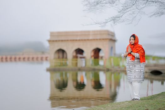 A woman in vibrant traditional attire stands in prayer beside a scenic lake with a historic structure in the background.