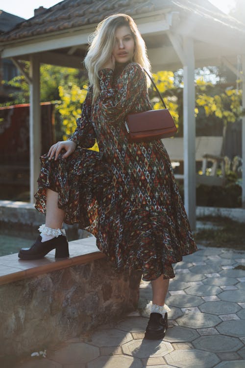 Full body of blond female in trendy socks and shoes with shoulder bag touching face while standing leaned on one leg and looking at camera in back lit in sunshine