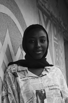 Black and white low angle of young smiling African American female in shawl on head and t shirt with text pattern standing leaned on concrete wall and looking at camera