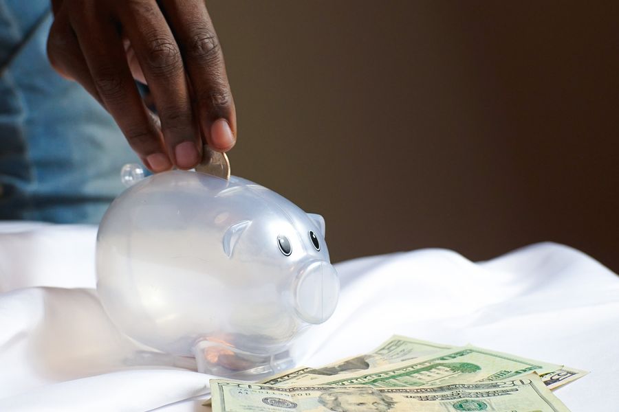 A hand dropping a coin into a piggy bank beside folded banknotes