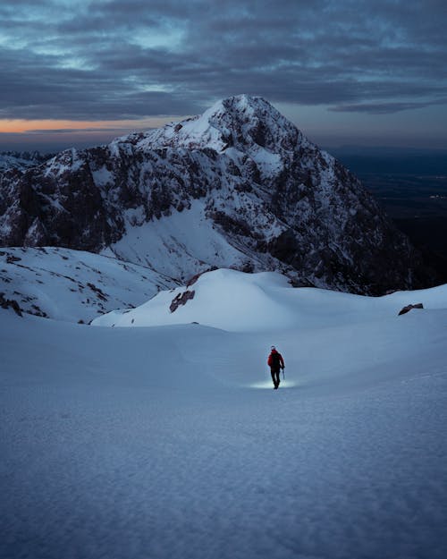 Gratis Orang Dengan Jaket Merah Dan Celana Hitam Berjalan Di Tanah Tertutup Salju Dekat Gunung Foto Stok
