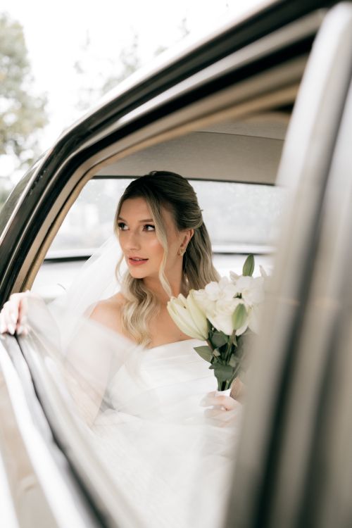Free Elegant bride in wedding dress holding a white lily bouquet, looking out from car window. Stock Photo