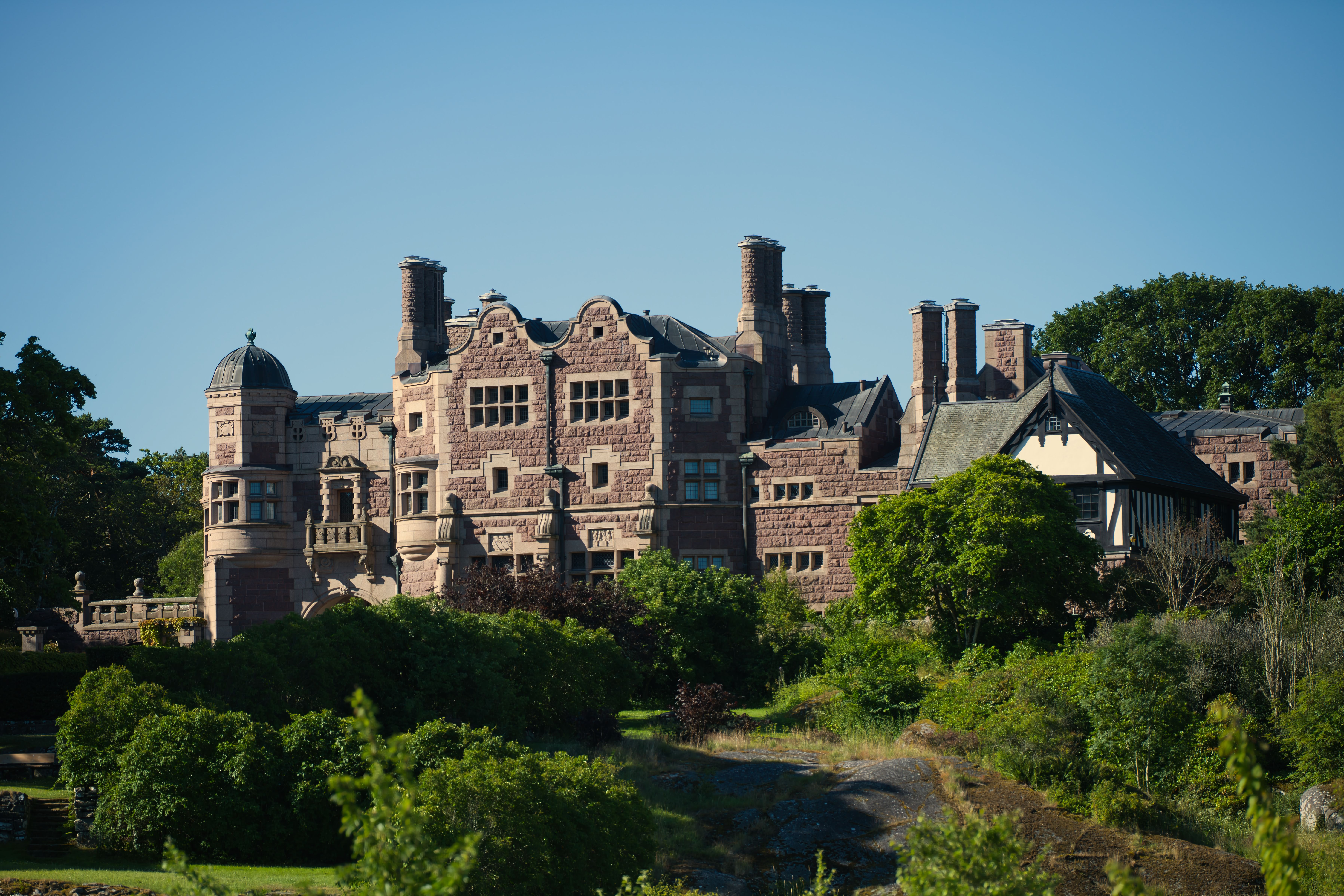 Elegant Historic Stone Mansion Nestled Among Vibrant Greenery And Clear Sky.