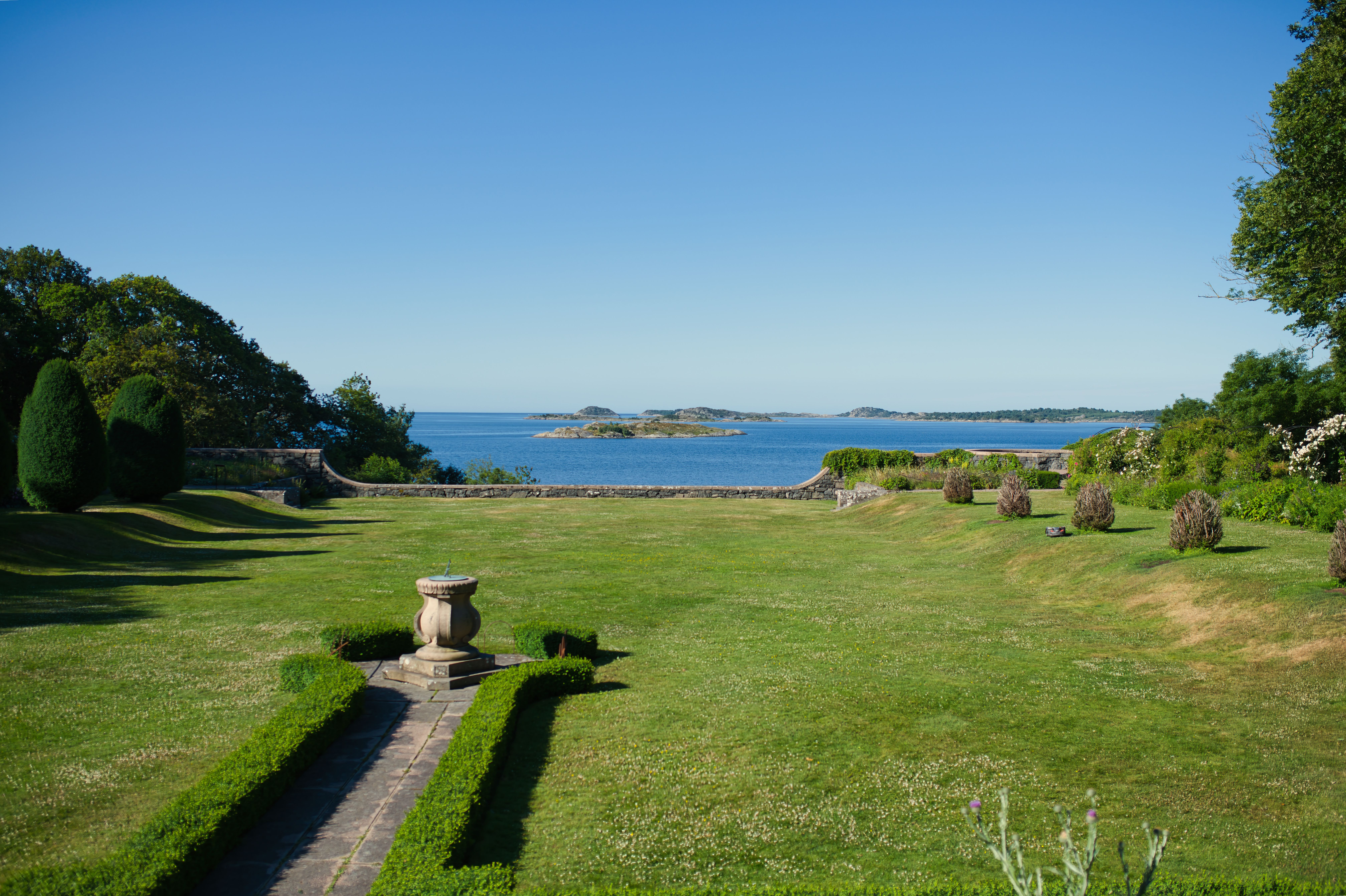 A peaceful coastal garden with lush greenery overlooking a calm sea under a clear blue sky.