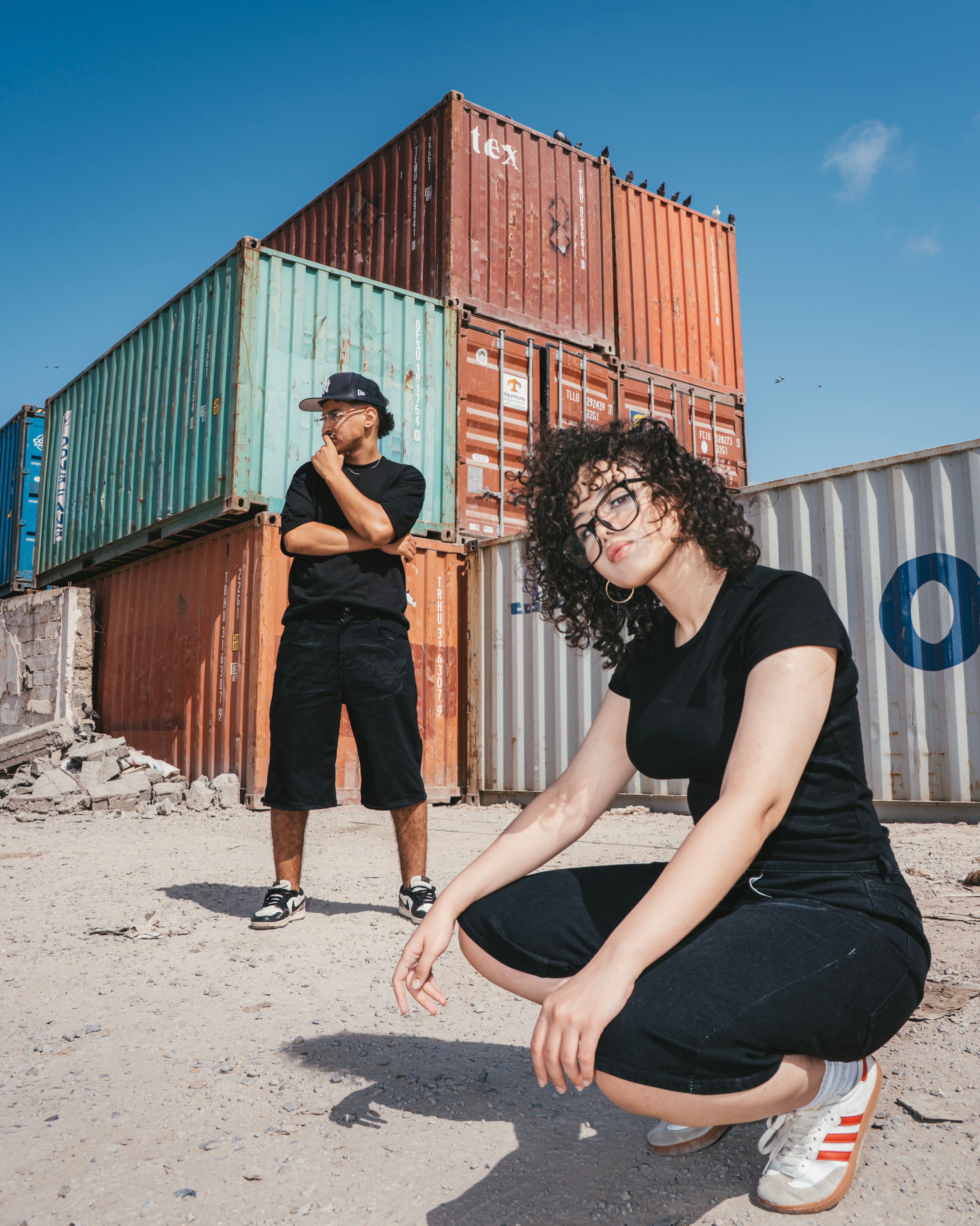 Two adults in casual black clothing pose around colorful shipping containers under a clear blue sky.